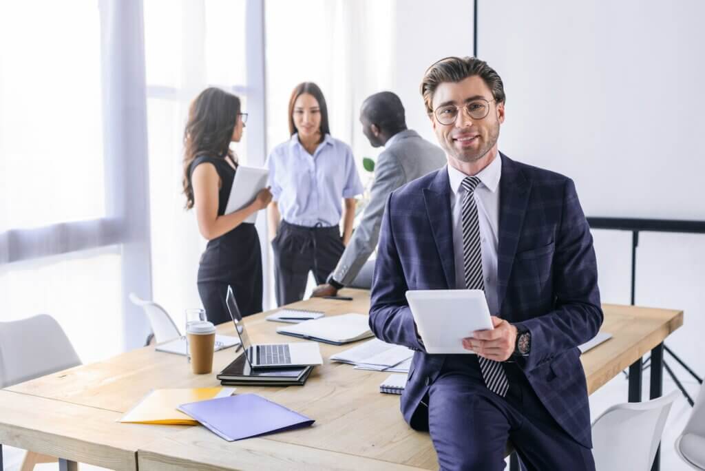 selective focus of caucasian businessman with tablet and multiracial colleagues behind in office
